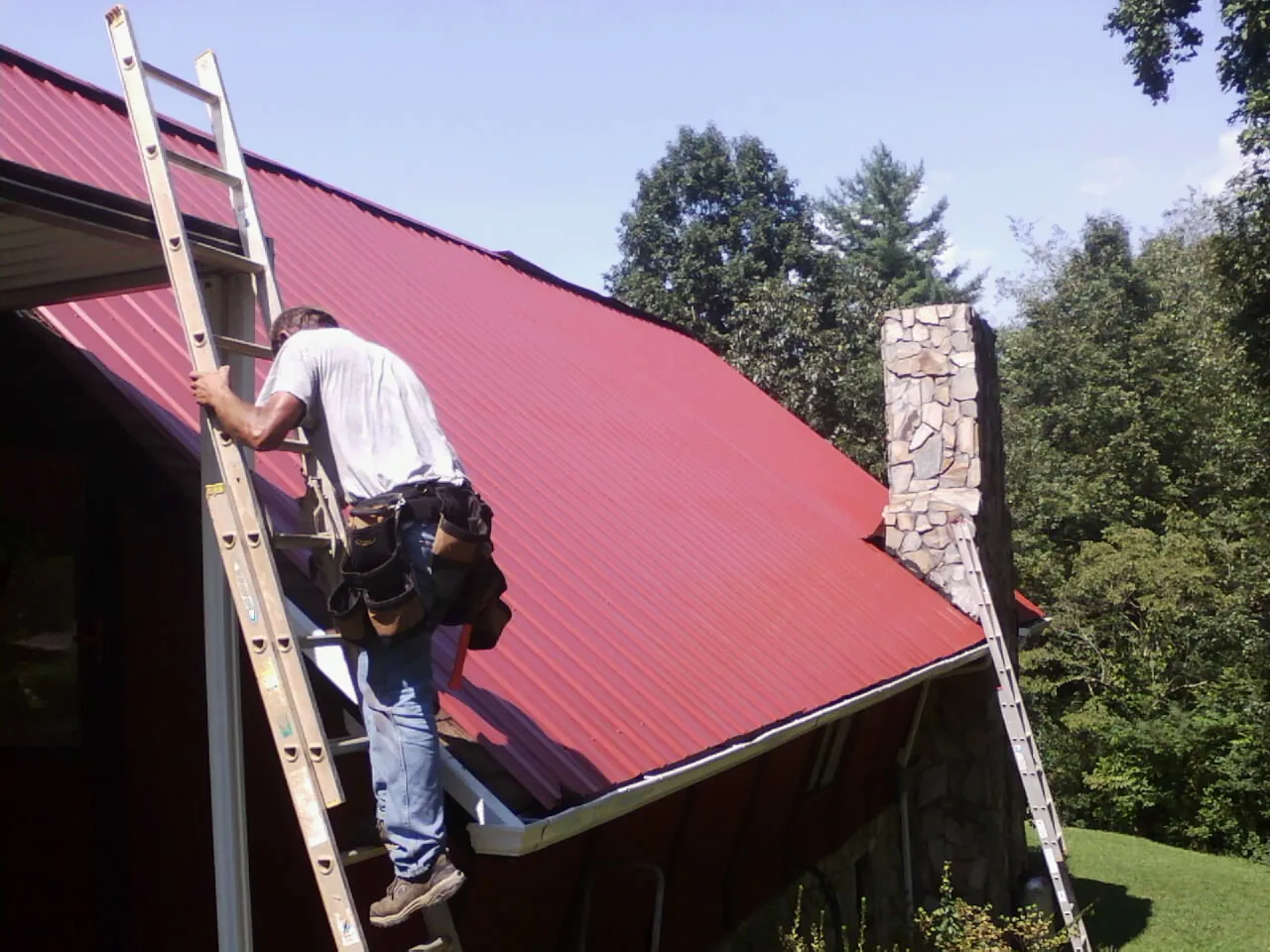 Roofer on a ladder beside a red metal roof and stone chimney — Soffit & Fascia Repair by Roofing Brothers in Richmondville